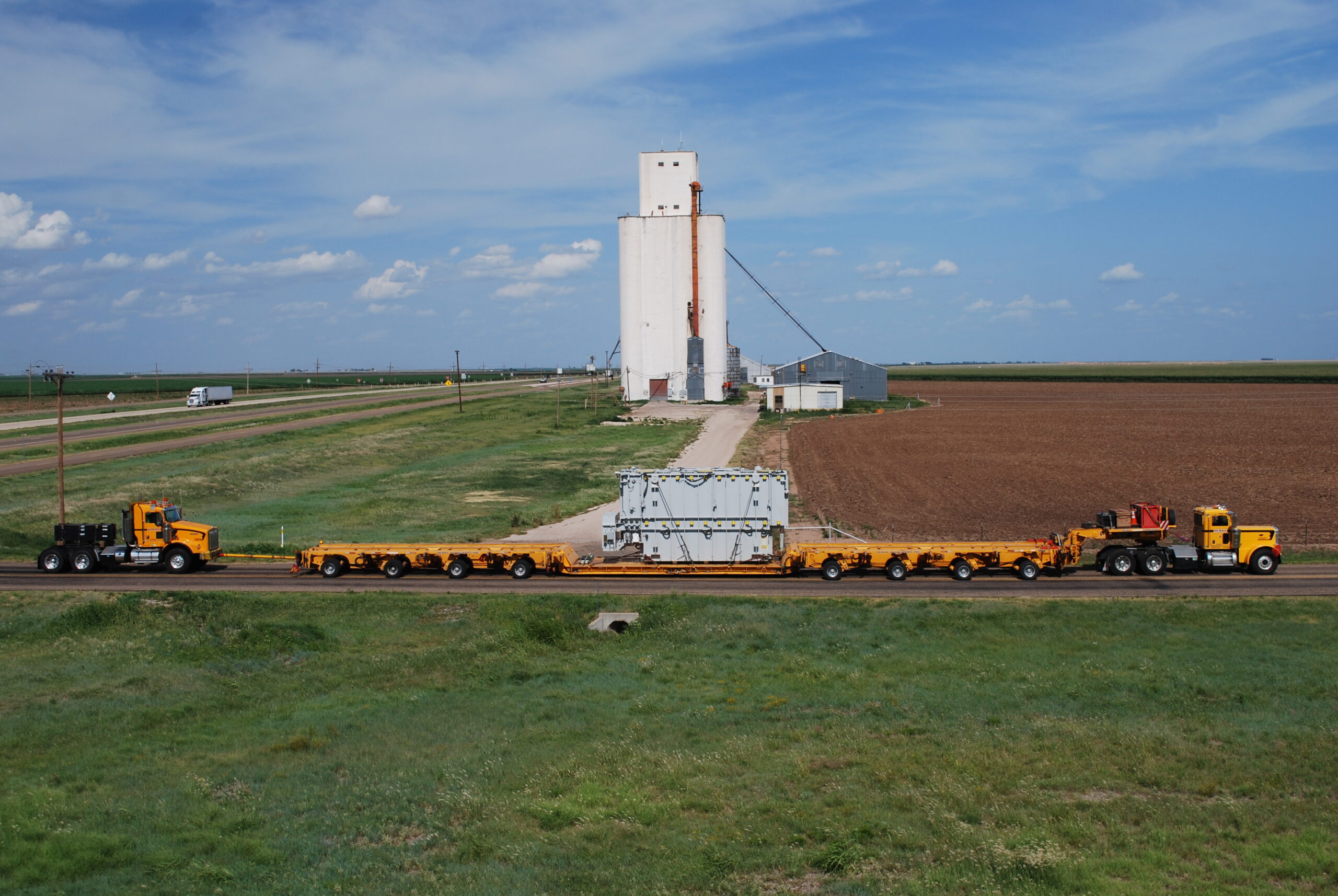 Aerial view of a long truck transporting a wind turbine blade, turning at a rural intersection surrounded by fields, grass, and a few cars on the road.