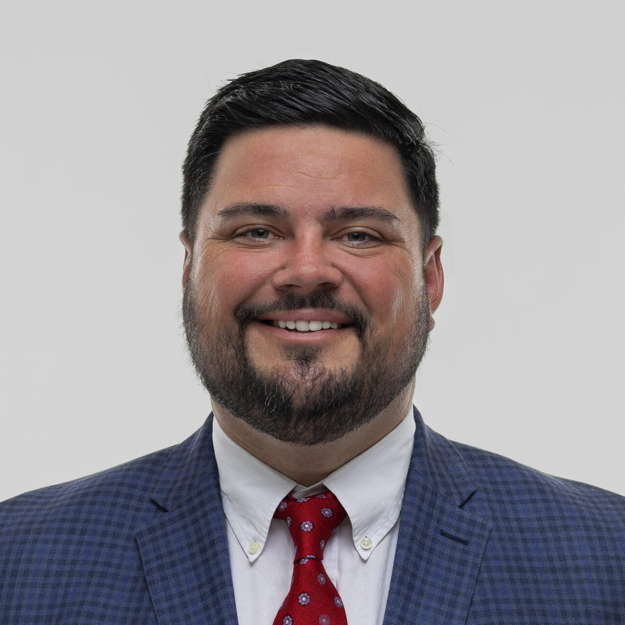Paul Gaspar in a dark suit, white shirt, and red tie smiles at the camera against a plain light background.