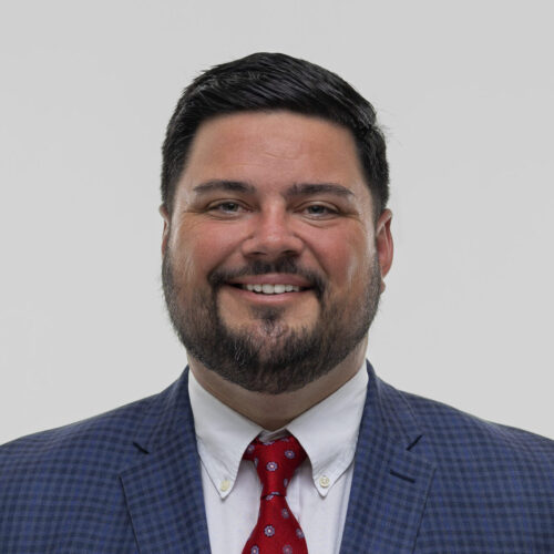 Paul Gaspar in a dark suit, white shirt, and red tie smiles at the camera against a plain light background.